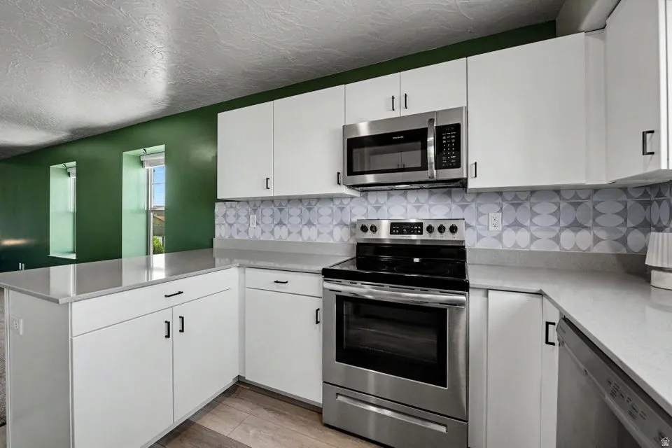 White cabinetry with black hardware, stainless steel appliances, patterned tile backsplash, light-toned countertops, and wood-finish flooring