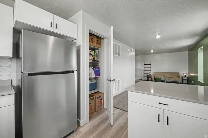 Stainless steel refrigerator, white cabinetry, light wood-finish flooring, light-colored countertops, and pantry storage