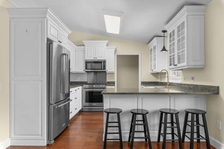 White cabinetry, stainless steel appliances, dark stone countertops, wood-finish flooring, and a breakfast bar