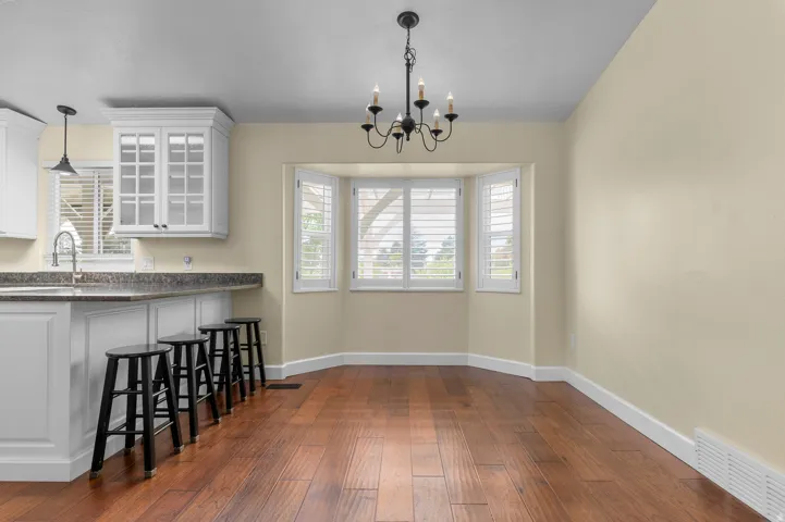 Wood-finish flooring, bay window with plantation shutters, kitchen island with built-in cabinetry, pendant lighting, and a chandelier
