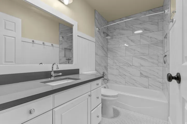 Vanity with undermount sink and dark countertop, chrome fixtures, white paneled cabinetry, white beadboard wainscoting, and white hexagon tile flooring