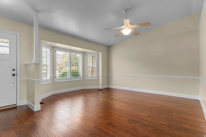 Wood-finish flooring throughout, bay window with plantation shutters, ceiling fan with light fixture, and crown molding accents