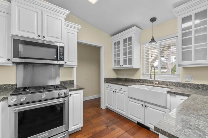 White cabinetry, stainless steel appliances, farmhouse sink, wood-finish flooring, and a window with blinds