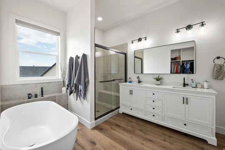 Freestanding soaking tub with dark-finish fixtures, a spacious double vanity with white cabinetry, wood-finish flooring, and a glass-enclosed shower