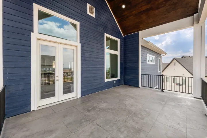 Expansive covered balcony featuring patterned concrete flooring, dark stained wood ceiling, white columns, black metal railings, and a double glass door entry with transom window