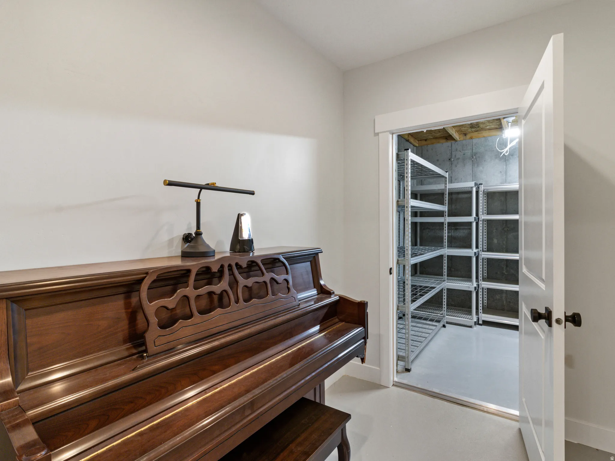 Music room featuring a wood-finish upright piano with decorative carved detailing, white painted walls, and direct access to a climate-controlled storage room with metal shelving and glass-panel door