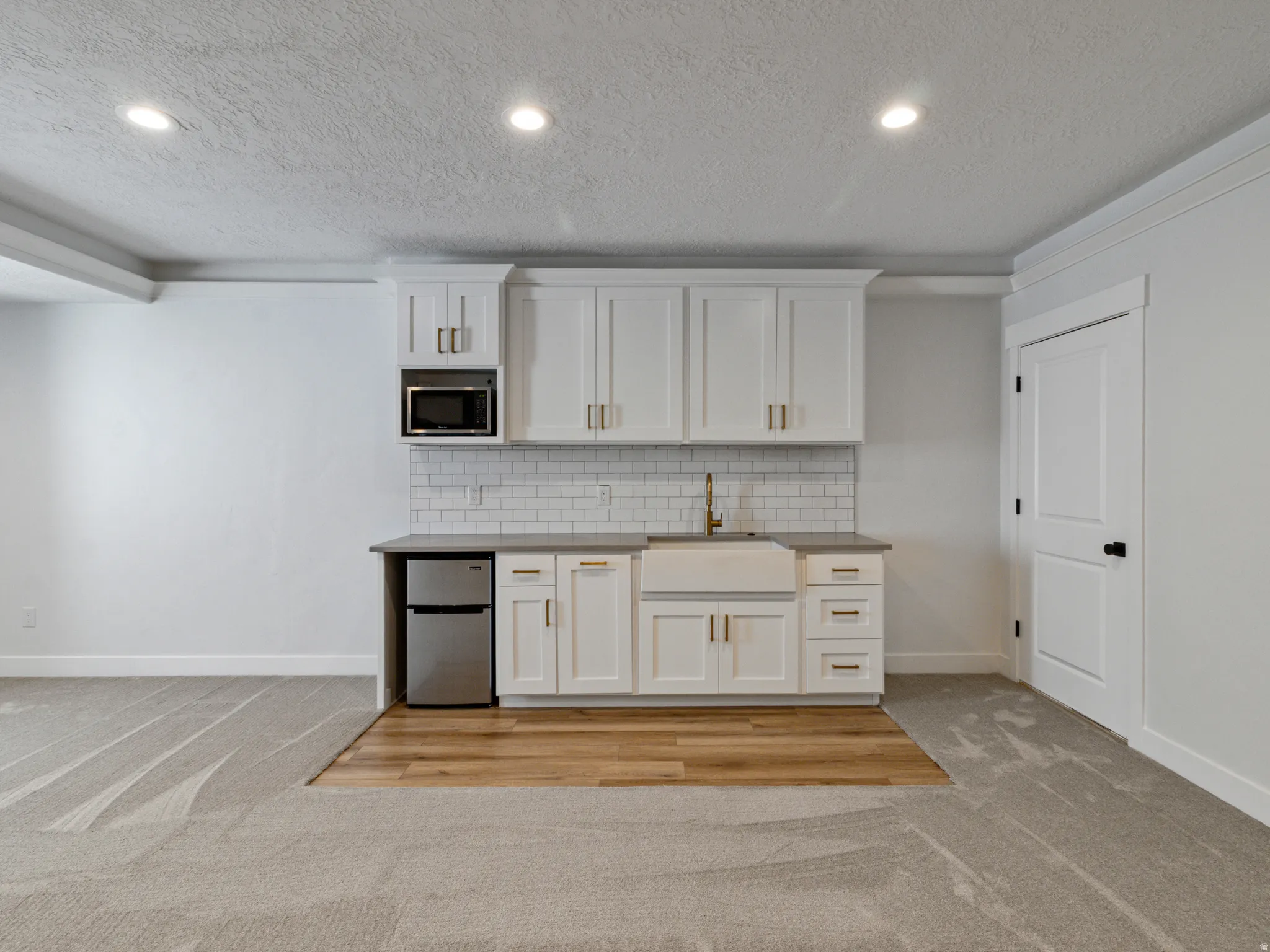 White shaker cabinetry, subway tile backsplash, farmhouse-style sink, stainless steel microwave, and wood-finish flooring