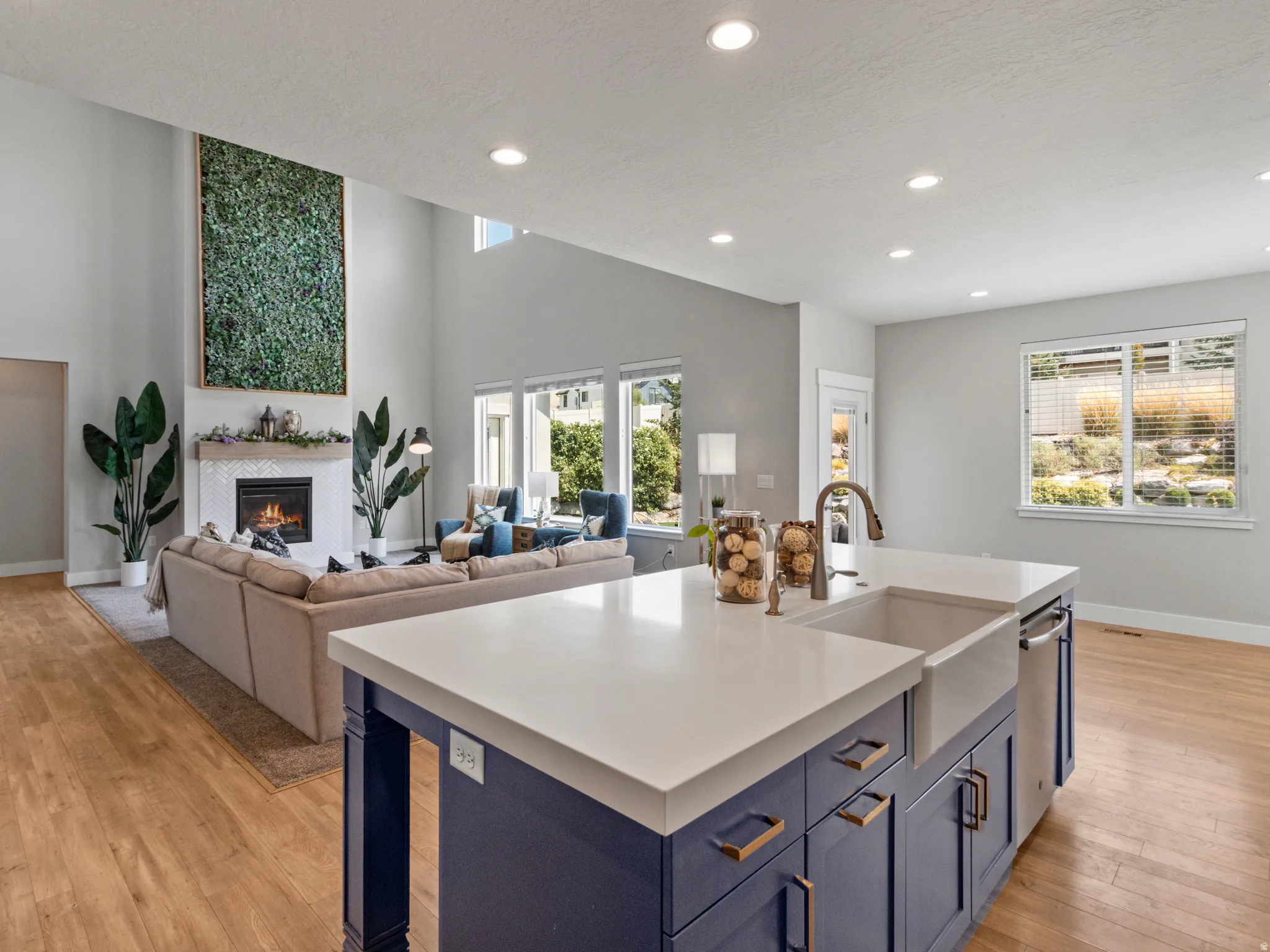 Open concept living space with wood-finish flooring, a kitchen island featuring a farmhouse sink and blue cabinetry, a fireplace with a white mantel, and recessed lighting
