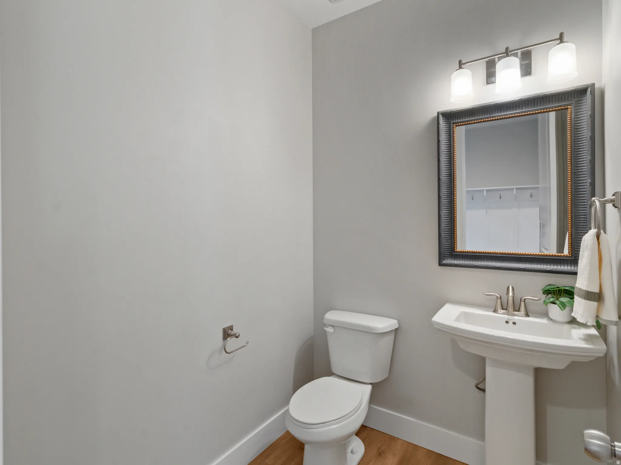 Pedestal sink with a brushed nickel faucet, framed mirror, three-light vanity fixture, and wood-finish flooring