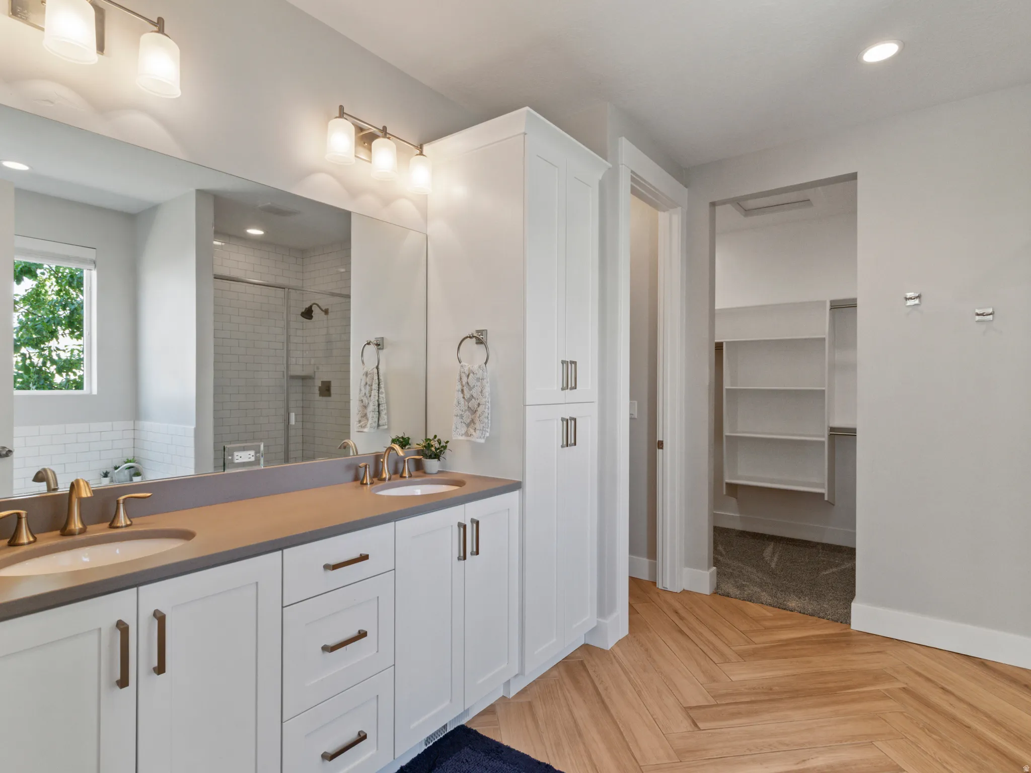 Primary bathroom featuring dual vanities with wood-finish countertops, white cabinetry with brushed metal hardware, an oversized framed mirror with integrated lighting, a walk-in shower with glass enclosure and subway tile surround, and direct access to a