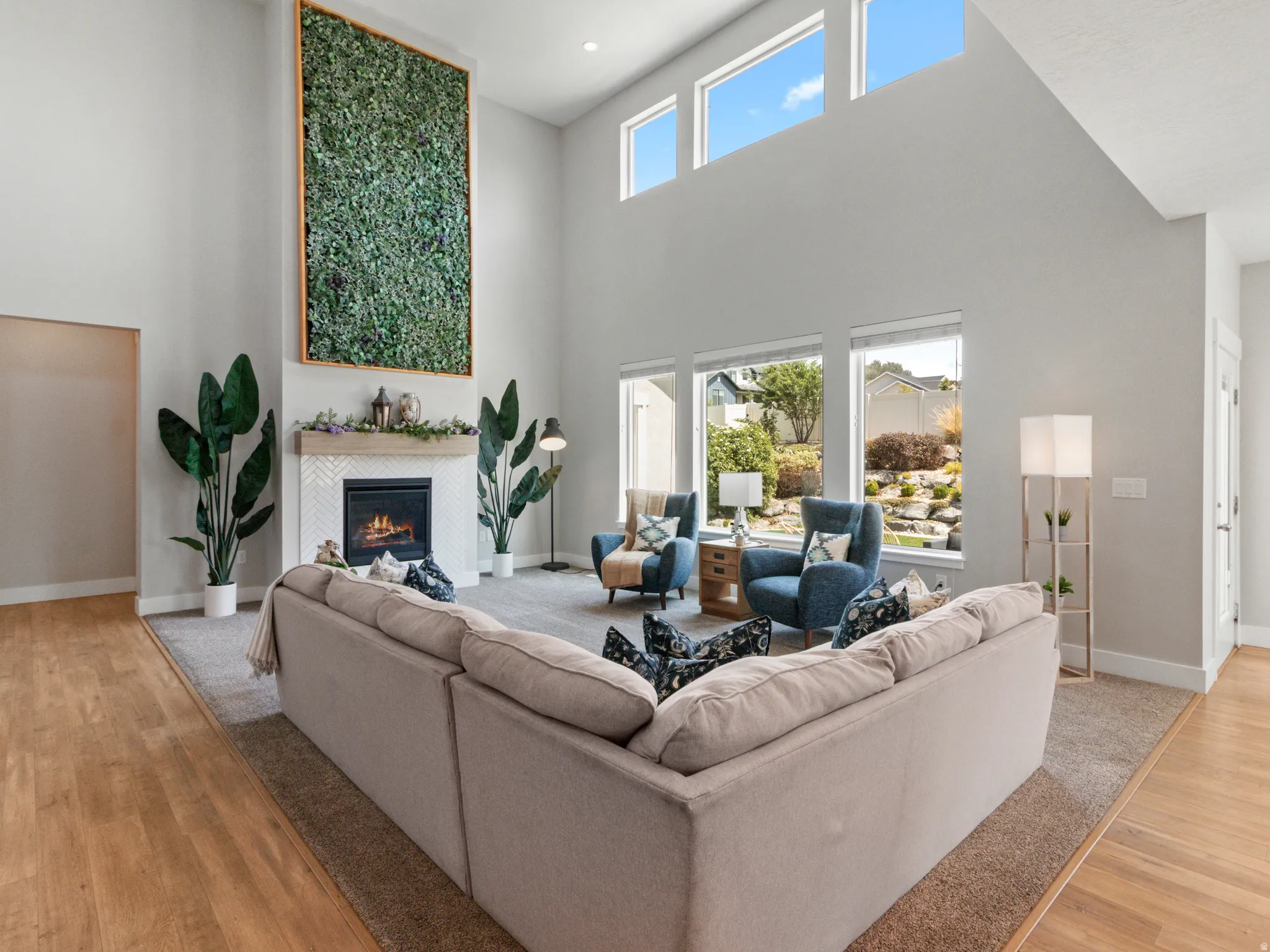 Soaring ceilings with transom windows, a fireplace featuring a herringbone-patterned tile surround and a wood mantel, expansive windows, and wood-finish flooring