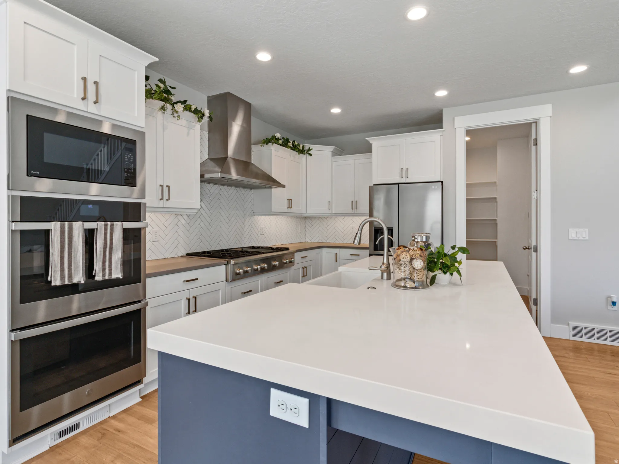 Modern kitchen featuring white shaker cabinetry, quartz countertops, a blue-painted island with waterfall edge, stainless steel appliances including dual wall ovens and a range hood, subway tile backsplash, recessed ceiling lighting, and wood-finish floor