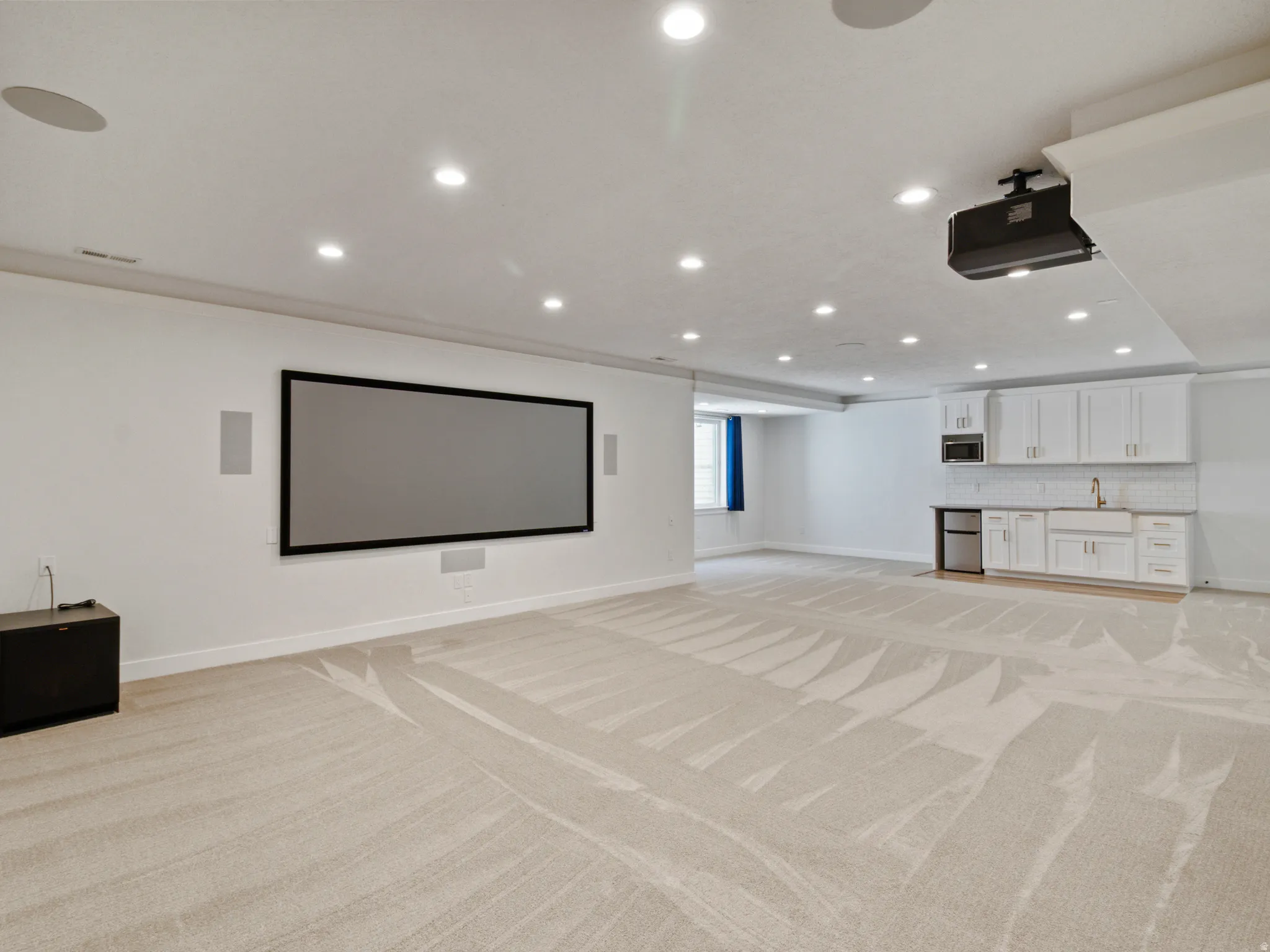 Expansive recreation room featuring recessed ceiling lighting, a framed projection screen, light wood-finish flooring with geometric patterning, and a built-in kitchenette with white cabinetry and stainless steel appliances