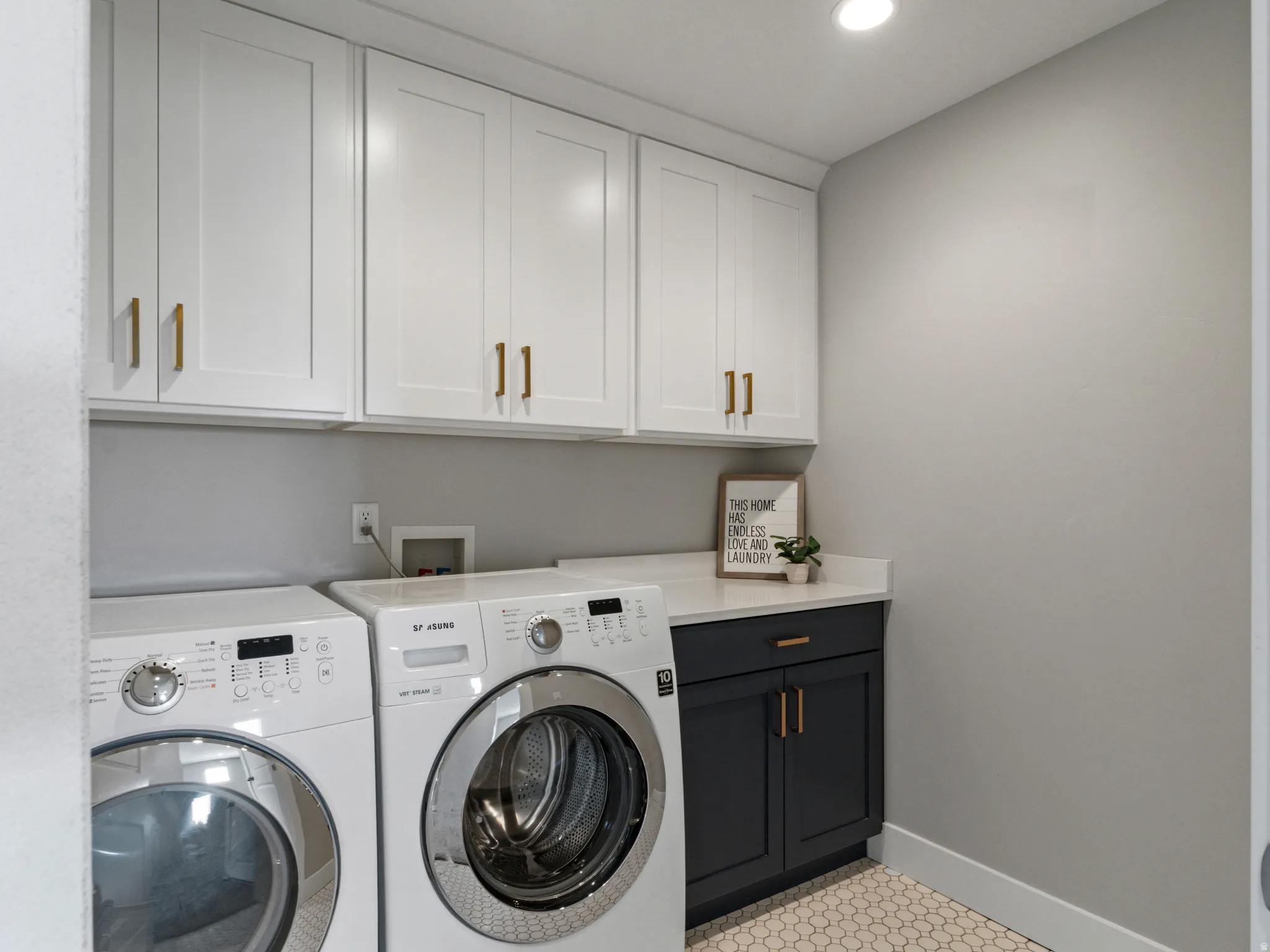 White shaker-style upper cabinetry with gold-finish hardware, dark lower cabinetry with gold-finish hardware, solid surface countertop, and hexagonal tile flooring