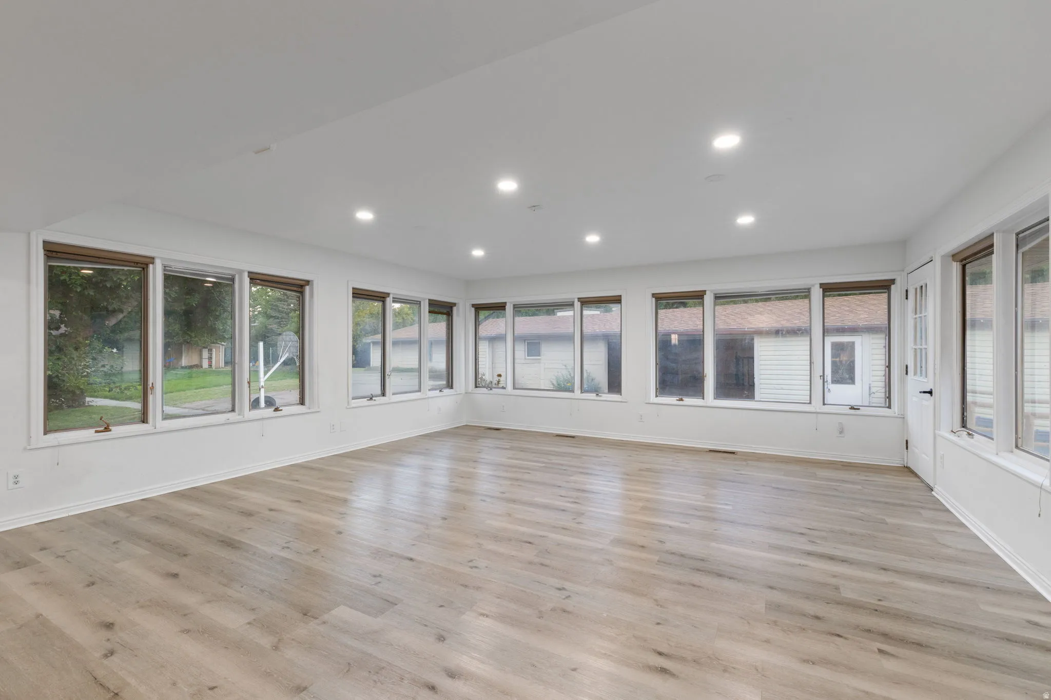 Spacious room featuring light wood-finish flooring, recessed lighting, multiple windows with natural wood trim, and a paneled door with a privacy glass insert