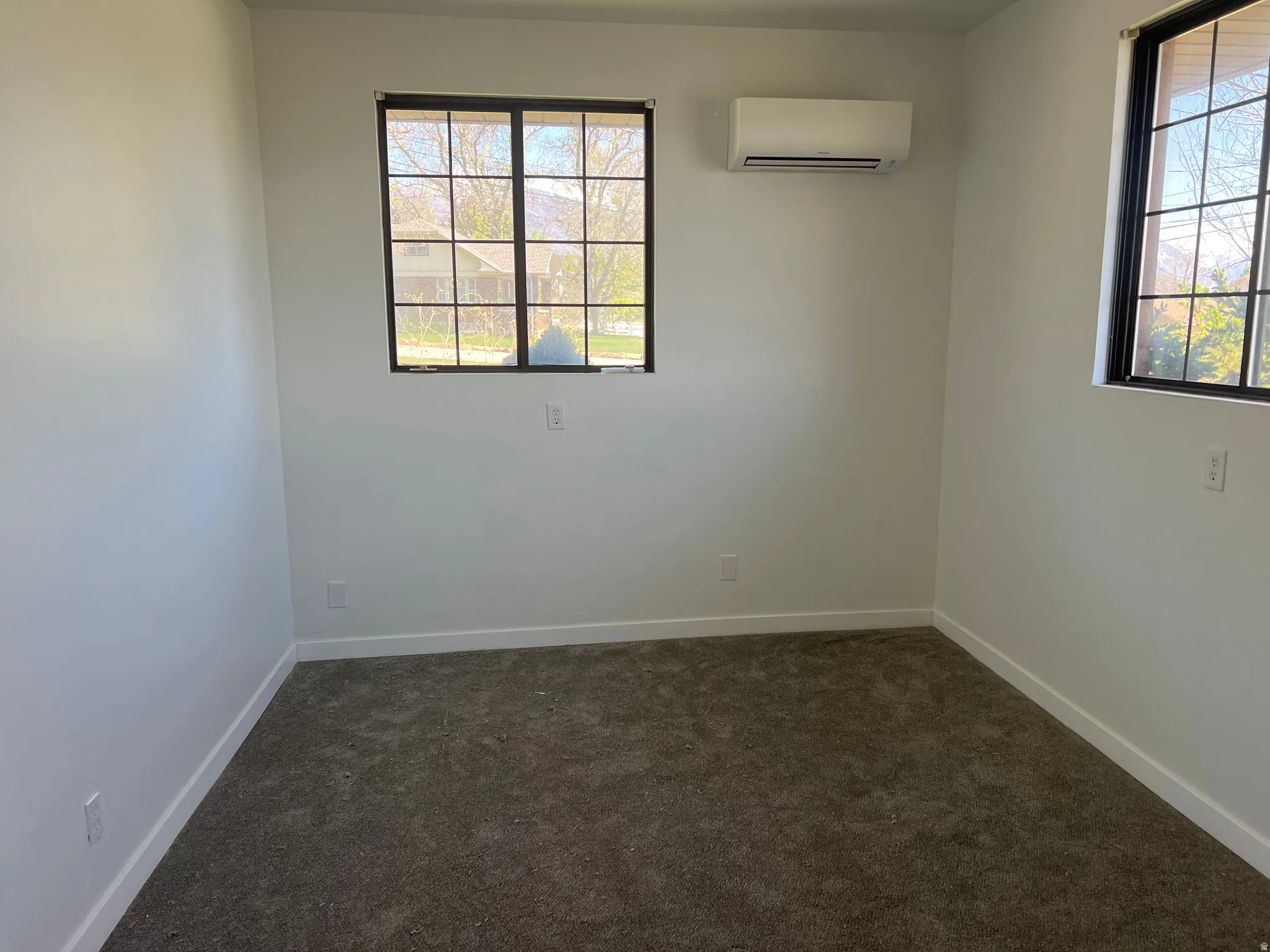 Neutral wall color, dark gray carpet, two windows with black frames and grids, and a wall-mounted mini-split HVAC unit