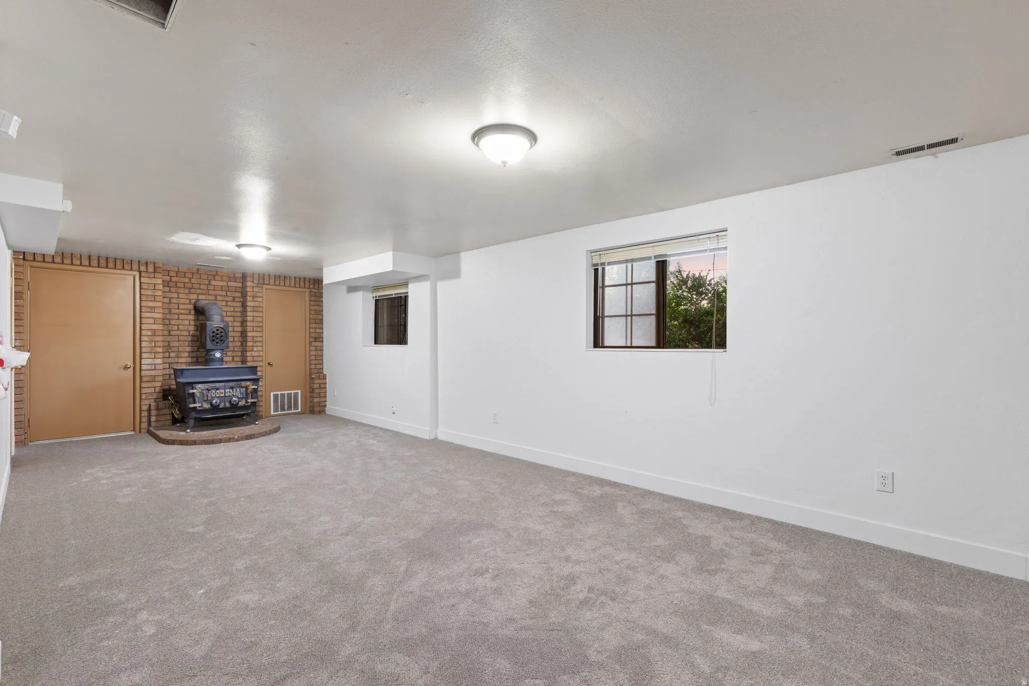Spacious room with light gray carpeting, a brick accent wall, and a prominent wood-burning stove on a raised hearth