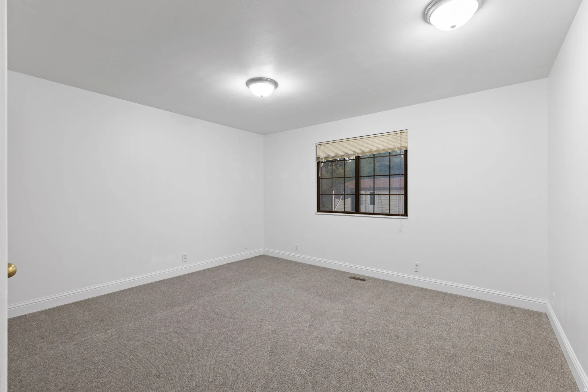 Neutral tone wall paint, light-colored carpeting, two ceiling-mounted light fixtures, window with blinds, and white trim