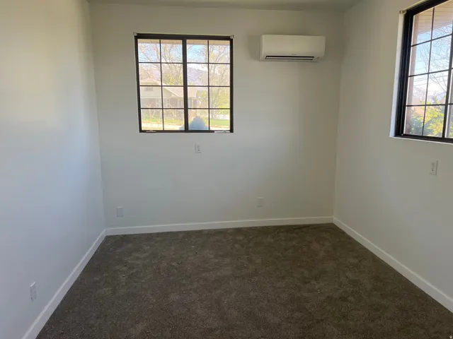 Neutral wall color, dark gray carpet, two windows with black frames and grids, and a wall-mounted mini-split HVAC unit