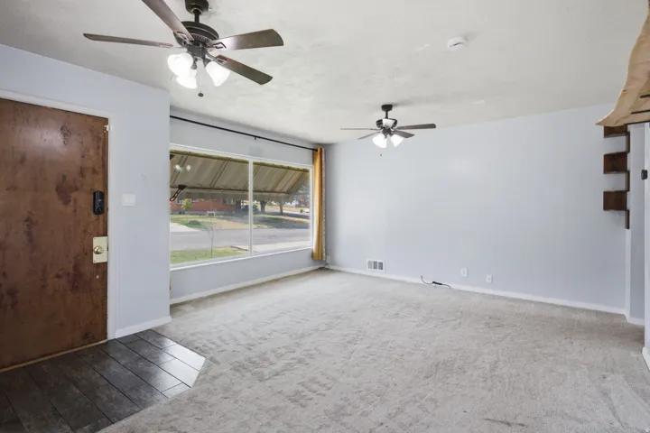 Spacious living area featuring neutral carpeting, dual ceiling fans with integrated lighting, large picture window with natural light, and dark wood-finish entry door