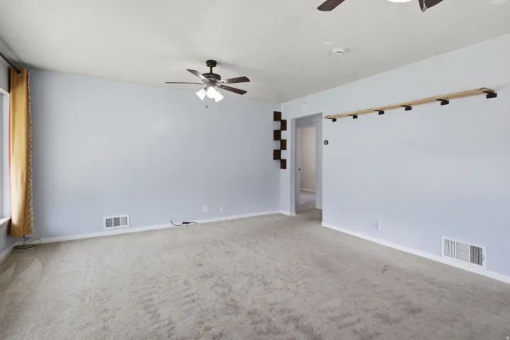 Light-colored carpeting, light blue wall paint, a window with curtains, and a ceiling fan with light fixture