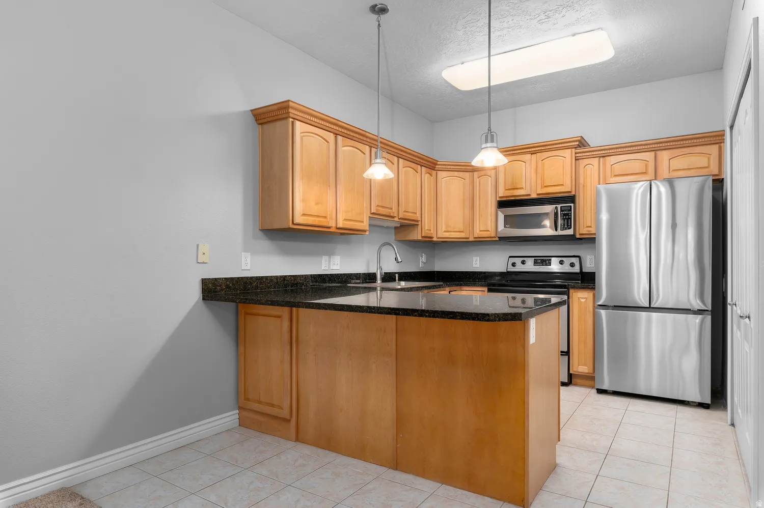 Kitchen featuring wood cabinetry, dark countertops, tile flooring, stainless steel appliances, and pendant lighting
