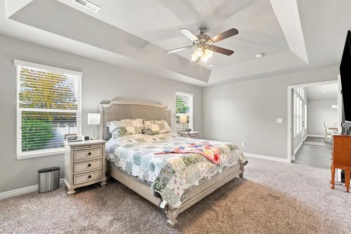 Tray ceiling with recessed lighting and ceiling fan, carpeted flooring, large window with blinds, light-toned wall paint, and an open doorway leading to another room