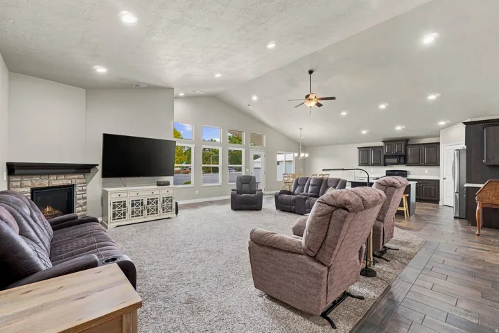 Vaulted ceiling with recessed lighting and a ceiling fan, stone-surround fireplace with mantel, window bank, and wood-finish flooring in the kitchen