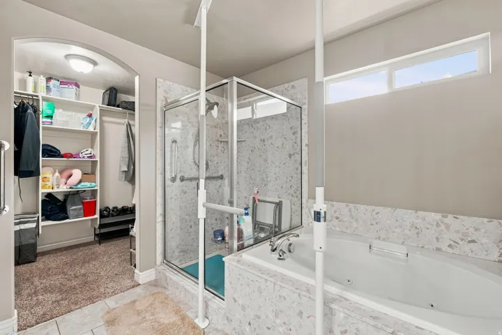Bathroom featuring a built-in tub, separate glass-enclosed shower, wall-mounted grab bars, and an arched entryway to a walk-in closet with built-in shelving