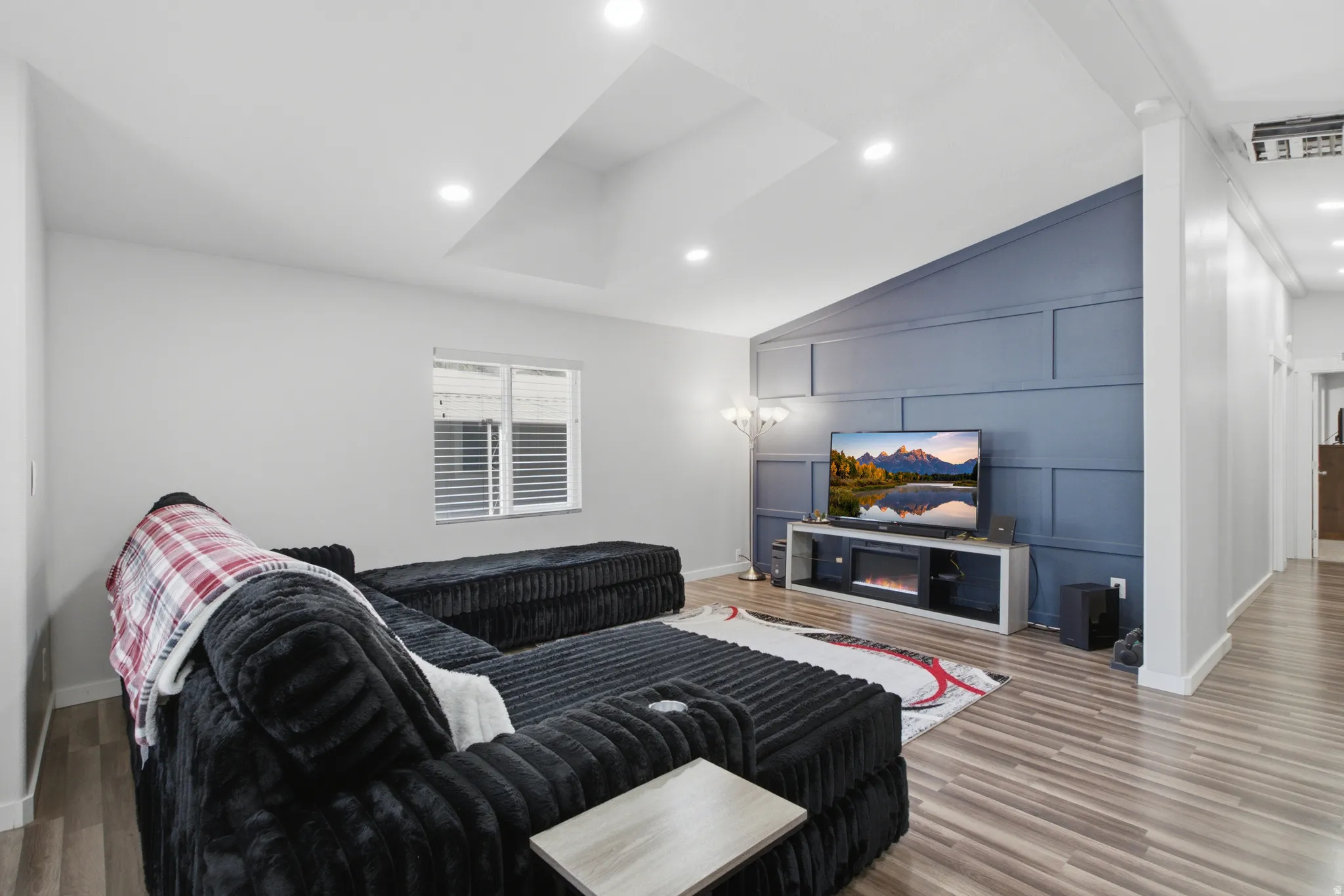 Vaulted ceiling with recessed lighting, accent wall with panel detailing, wood-finish flooring, and a window with blinds