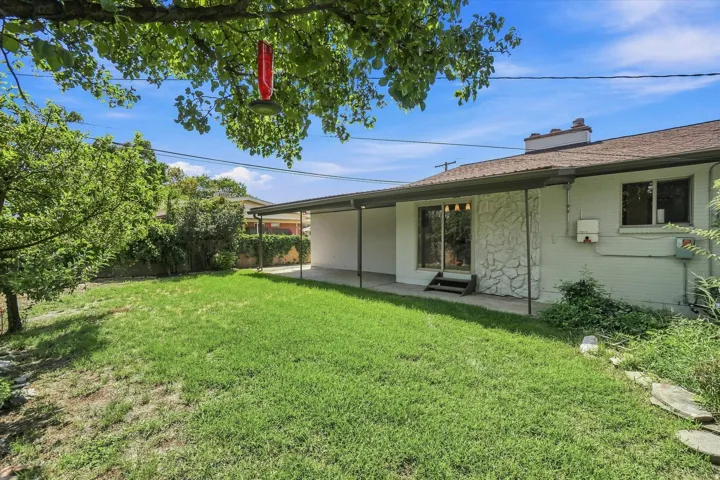Expansive rear yard with a covered patio, stone accent wall, and mature trees