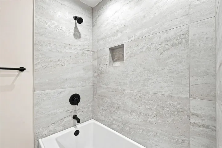 Bathtub with matte black fixtures, stone-look tile surround, and an integrated shower niche