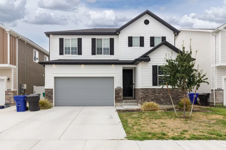 Two-story residence featuring white siding, black window shutters, a dark roof, stone-finish accents, and an attached garage with a gray door