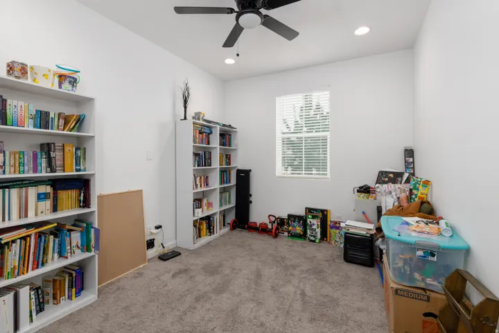 Bright, versatile room featuring built-in shelving with ample storage capacity, carpeted flooring, a ceiling fan with integrated lighting, and a large window with plantation shutters providing natural light
