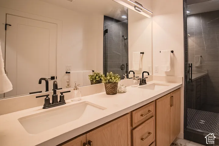 Dual undermount sinks with matte black fixtures, light-toned countertop, natural wood-finish cabinetry, frameless glass shower enclosure, and textured gray shower tile