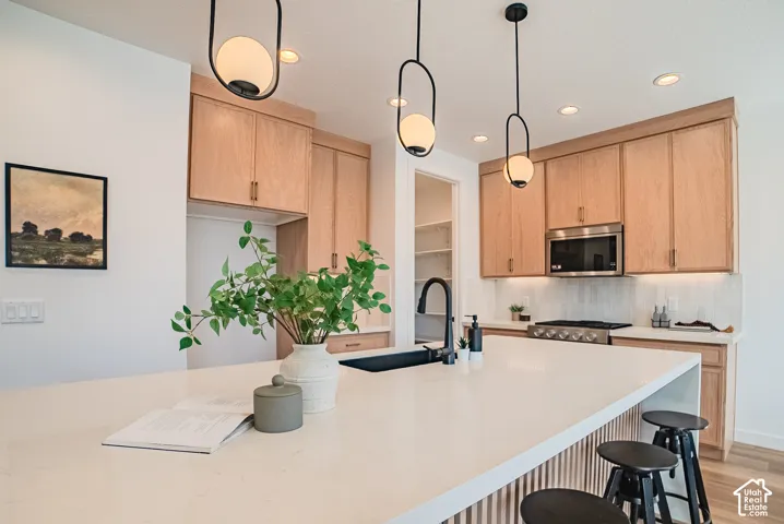 Kitchen featuring a large central island with seating, stainless steel appliances, light wood-finish cabinetry, under-cabinet lighting, and modern pendant fixtures