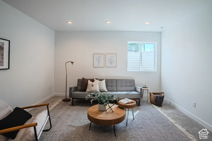 Recessed ceiling lighting illuminates the space, featuring white walls, a single window with pleated shades, and carpeted flooring
