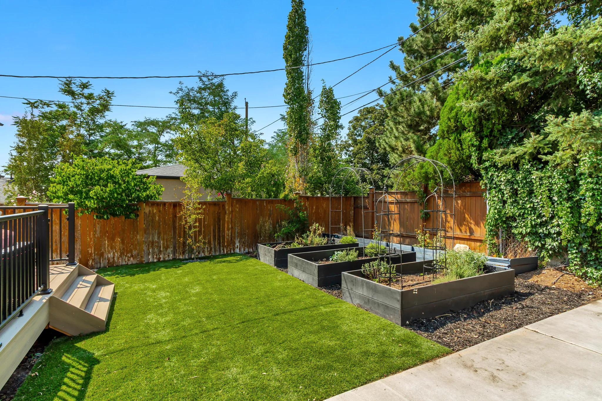 Outdoor space featuring a composite deck with black metal railings, artificial turf, multiple raised garden beds, and mature perimeter trees