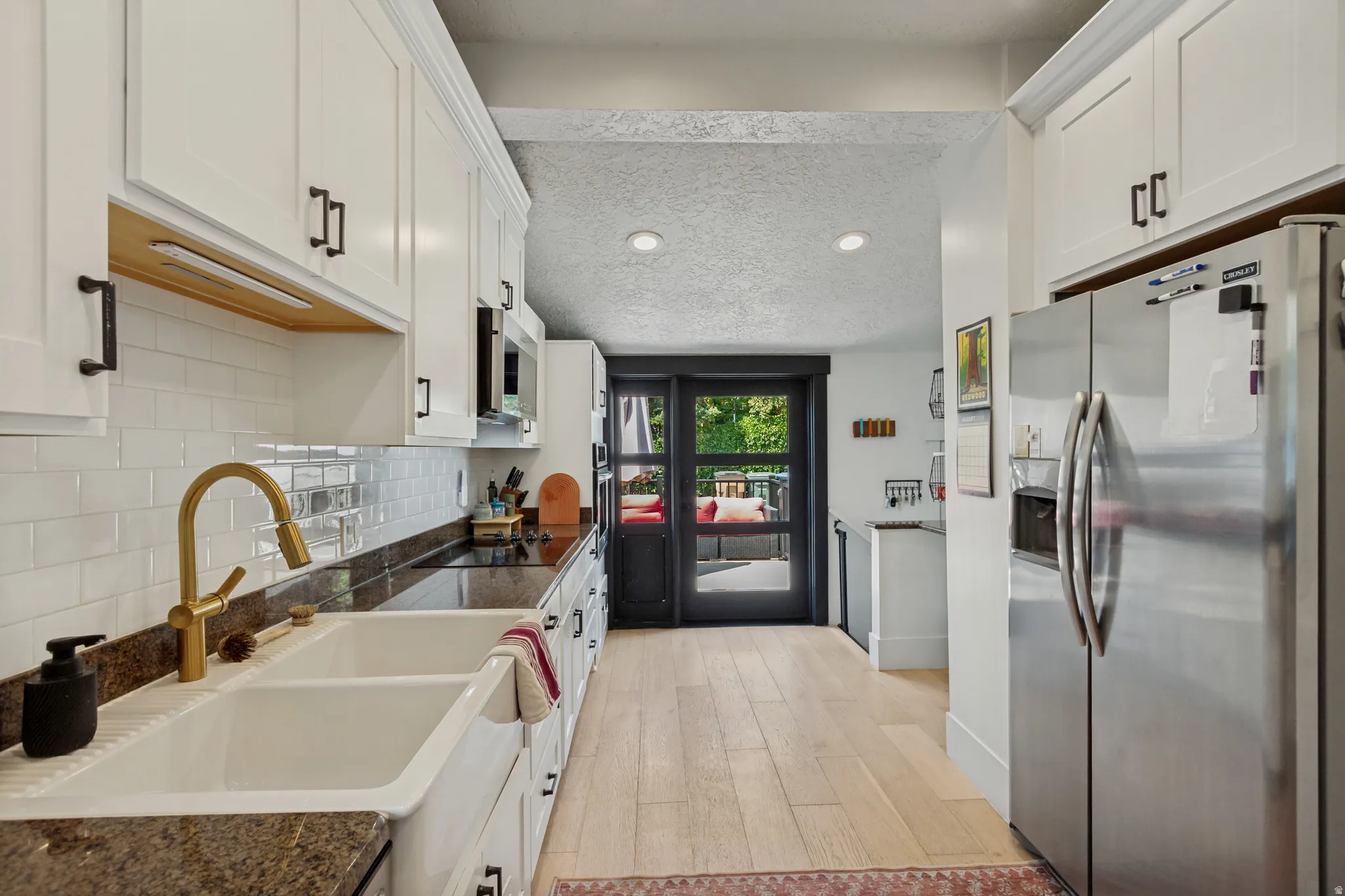 White shaker cabinetry with matte black hardware, white subway tile backsplash, a double basin farmhouse sink with a gold-finish faucet, and light wood-finish flooring