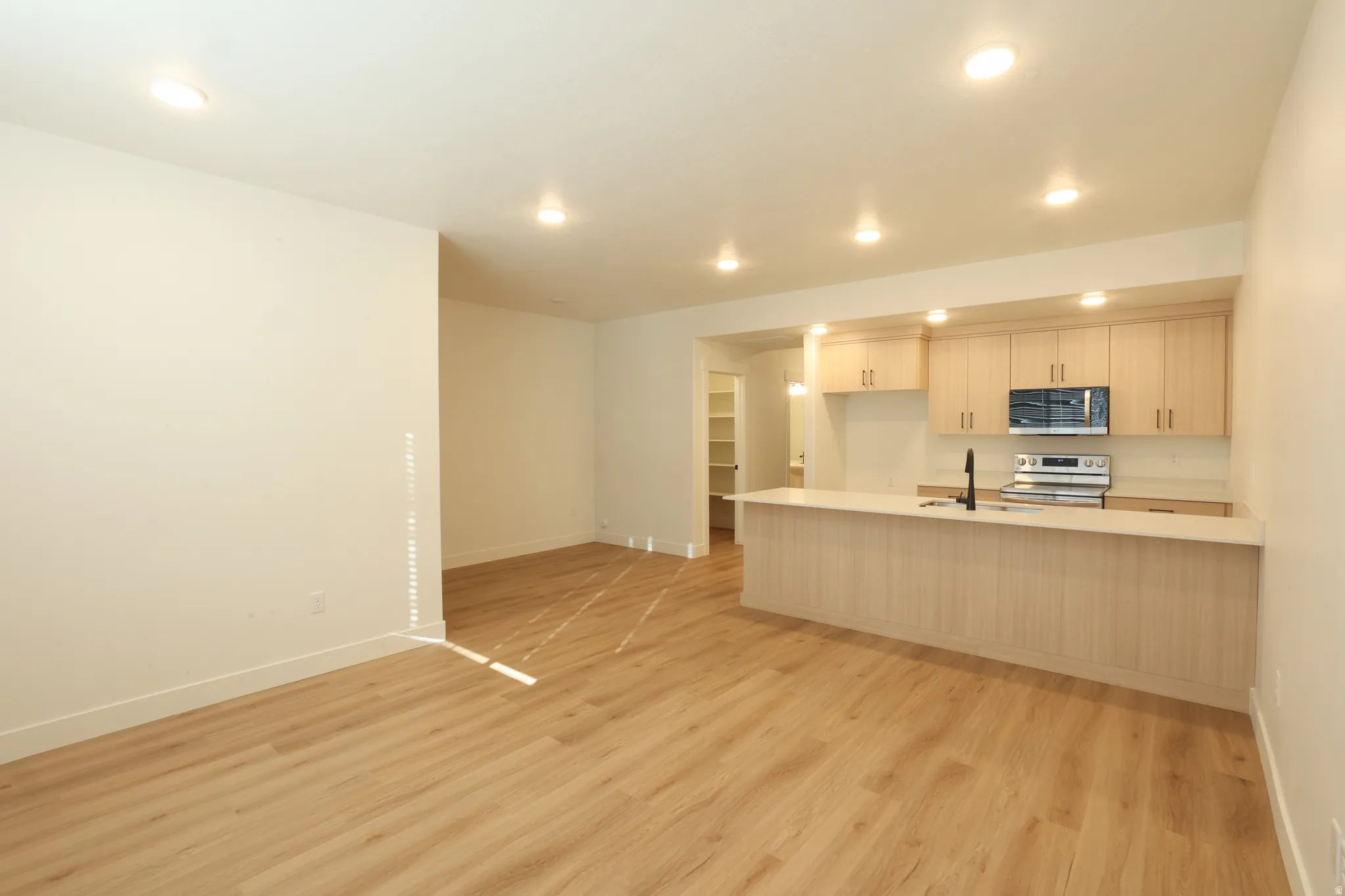 Open-concept living area featuring wood-finish flooring, recessed lighting, and a kitchen with light wood cabinetry, stainless steel appliances, and a breakfast bar with a light-toned countertop