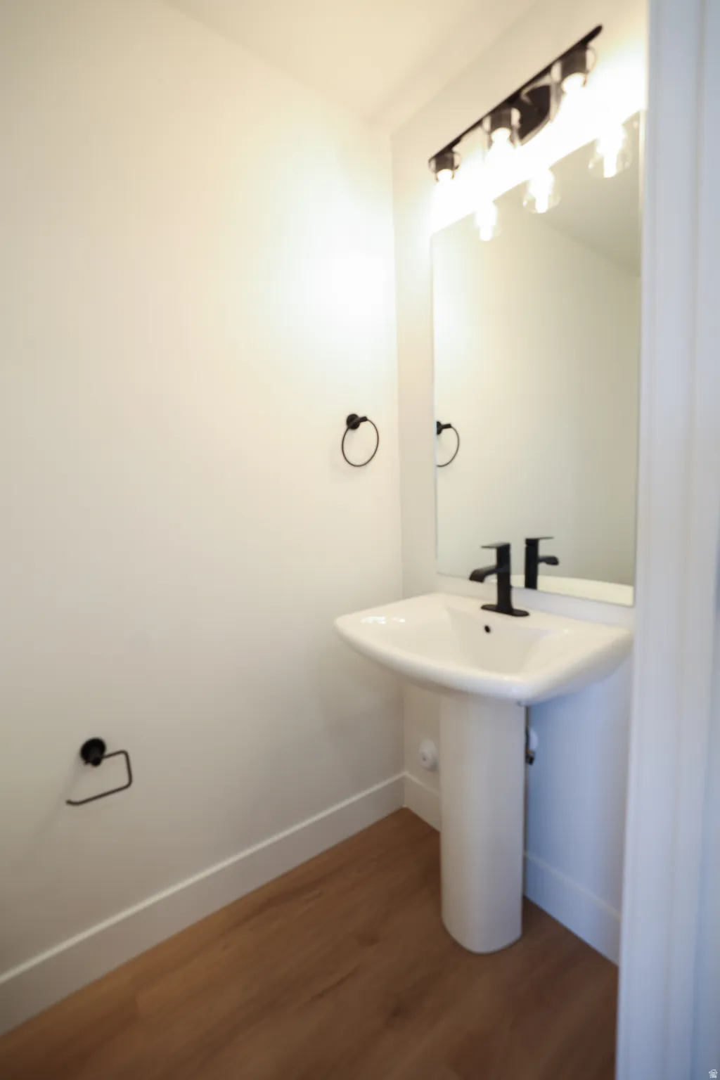 Pedestal sink with matte black faucet, wall-mounted mirror with vanity lighting, and wood-finish flooring