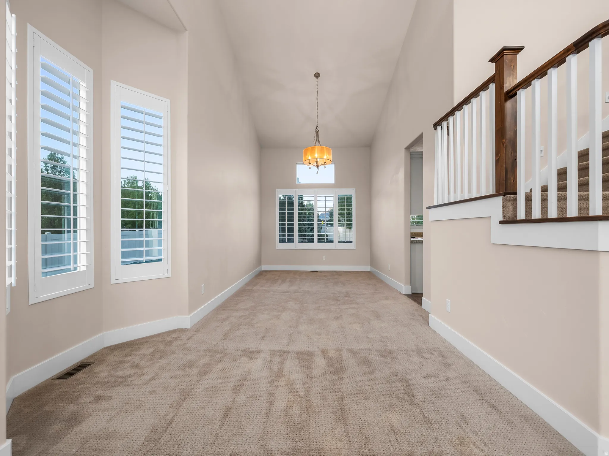 Vaulted ceiling with an amber-toned chandelier, multiple windows with white plantation shutters, and a staircase featuring a wood-finish handrail and white balusters