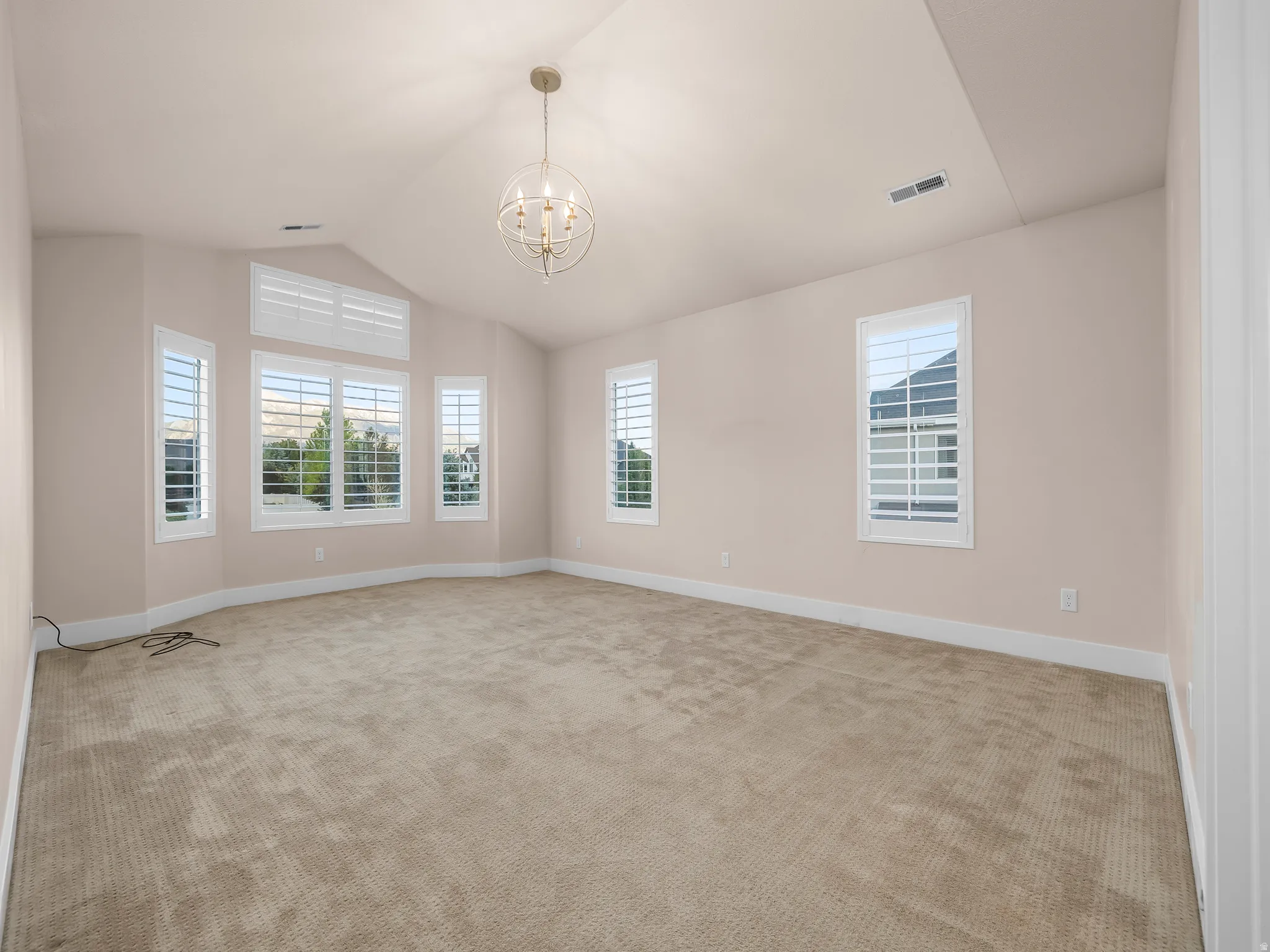 Vaulted ceiling with an orb chandelier, multiple windows with plantation shutters, light-toned carpeting, and neutral wall tones