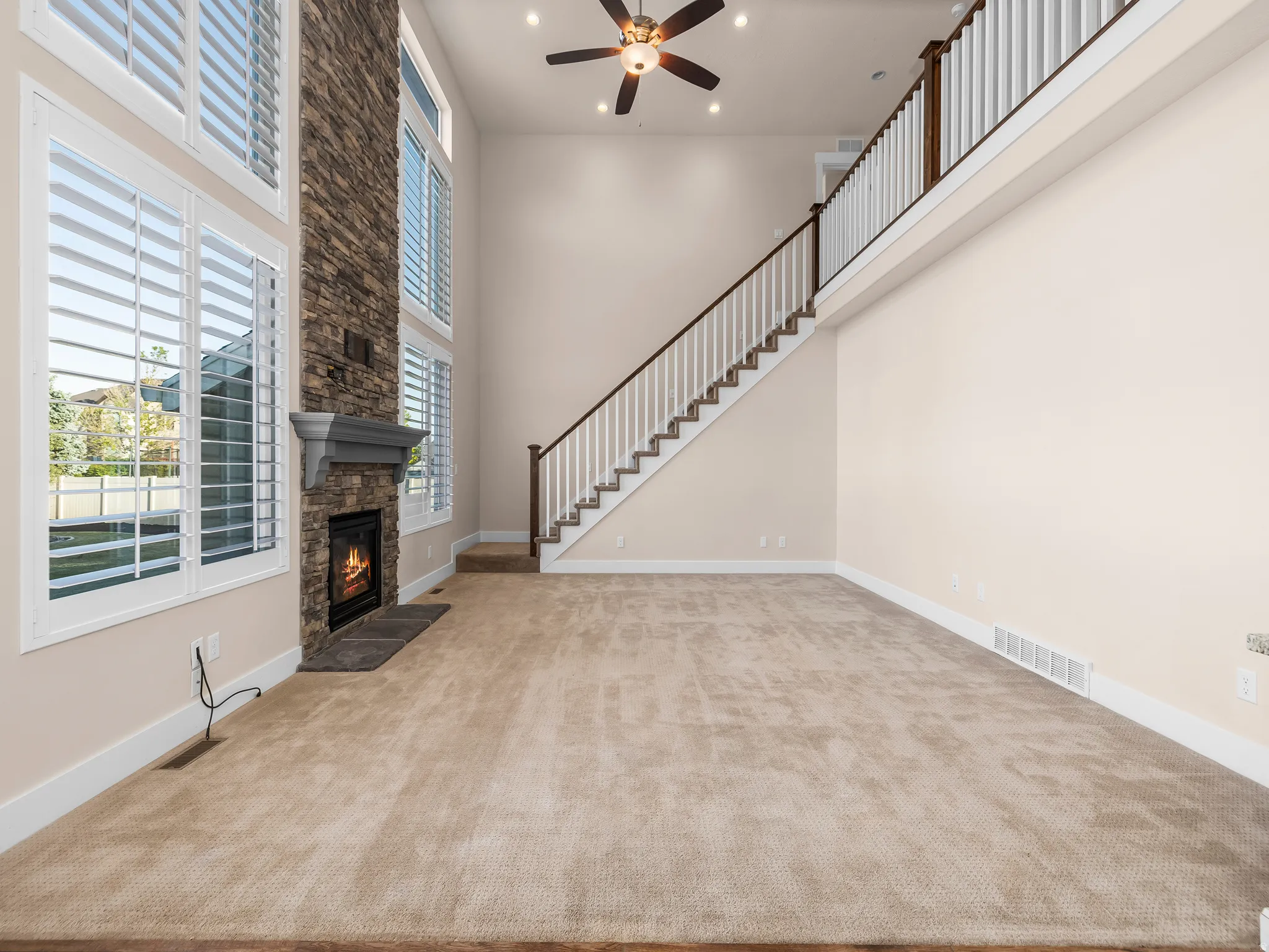 Two-story living area with a stone-clad fireplace and mantel, tall windows with plantation shutters, a multi-tiered staircase with wood handrails, and a ceiling fan with integrated lighting