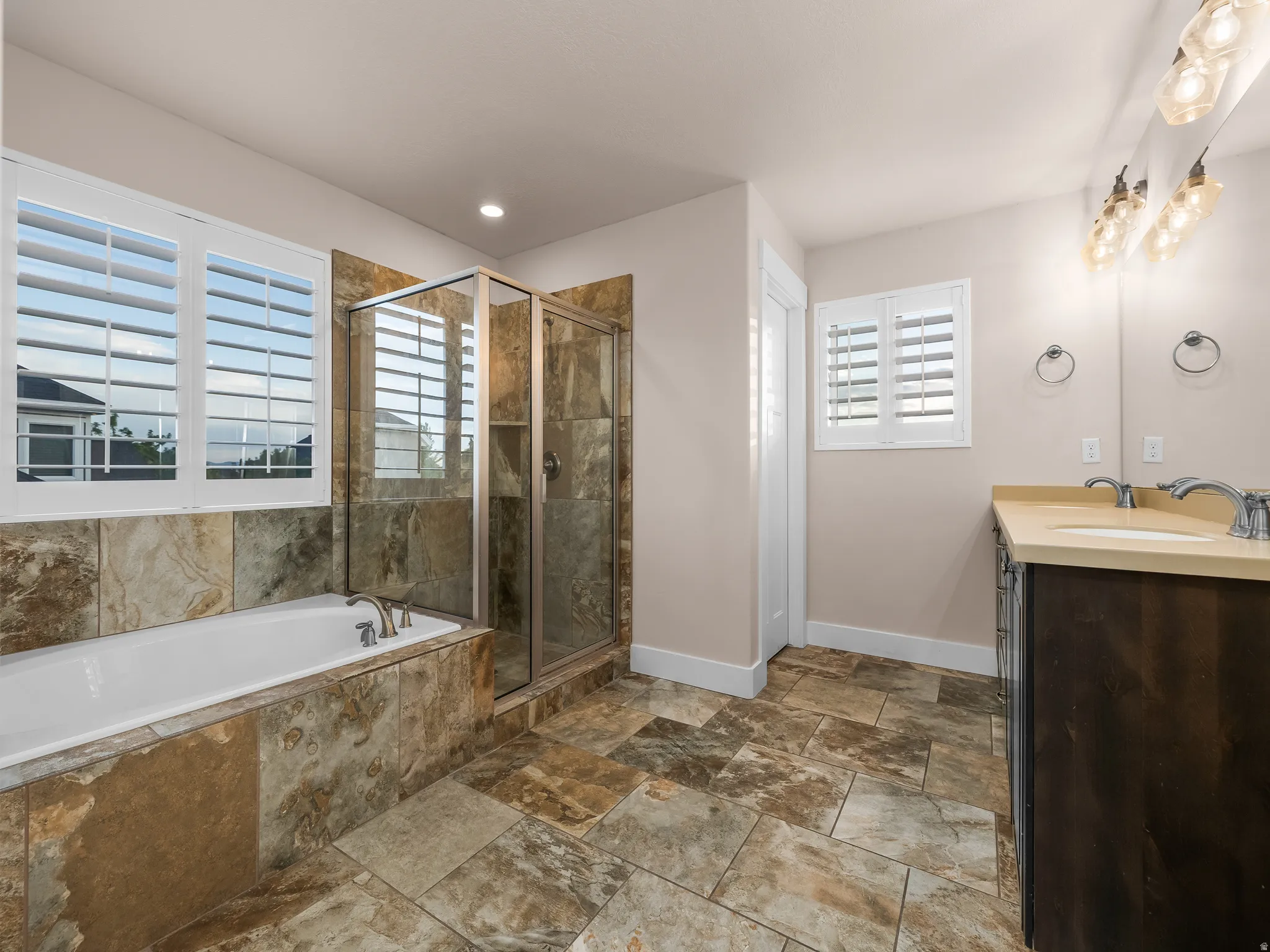 Tiled flooring and shower surround, glass-enclosed shower, built-in bathtub, dual vanity with solid surface countertop, and plantation shutters