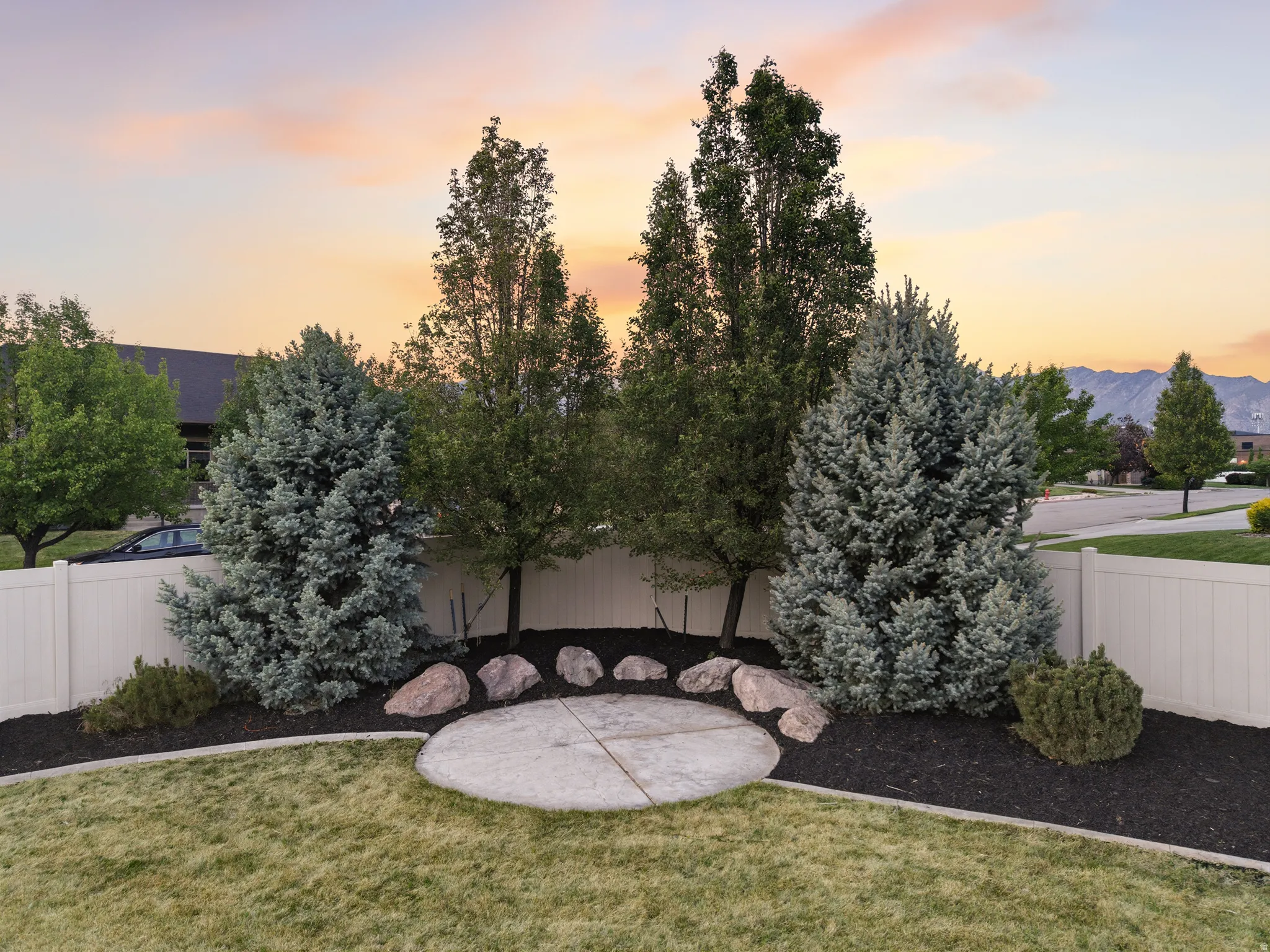 Expansive backyard featuring a manicured lawn, established trees, a white vinyl fence, a circular concrete patio, and decorative boulders