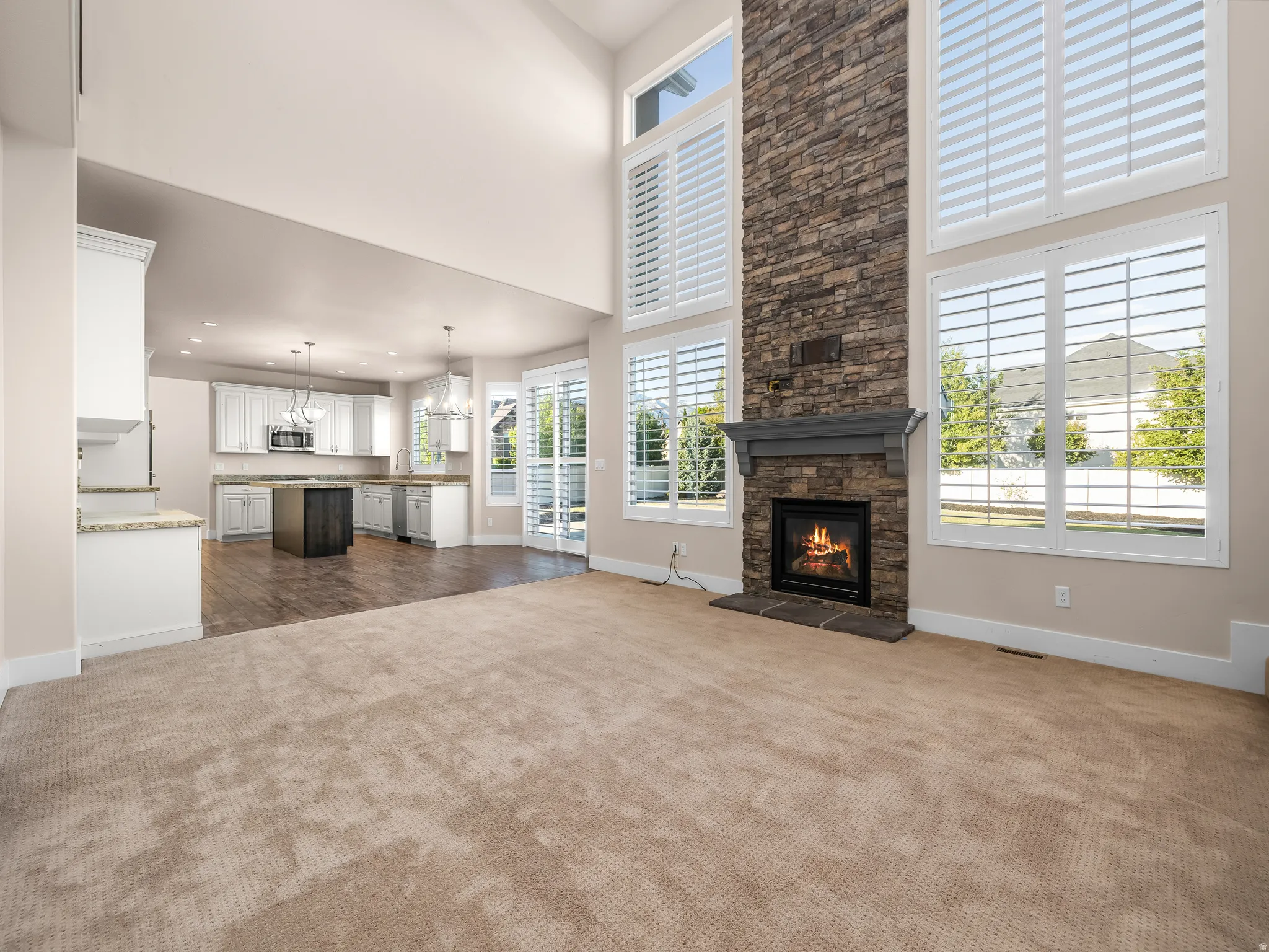 Two-story stone fireplace with mantel, white plantation shutters, vaulted ceilings, and a kitchen featuring white cabinetry, a center island, and built-in microwave