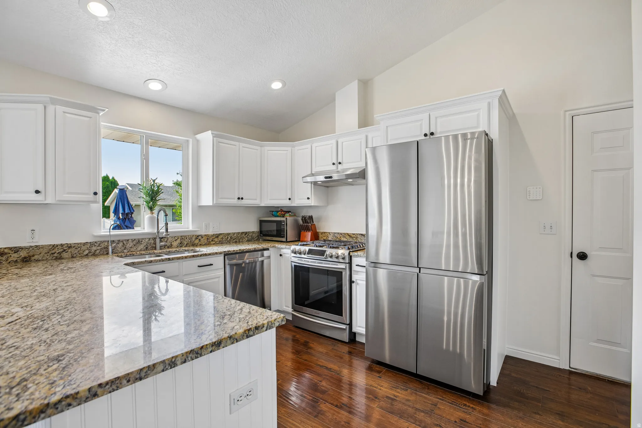 Kitchen featuring granite countertops, wood-finish flooring, white cabinetry, stainless steel appliances, and a vaulted ceiling
