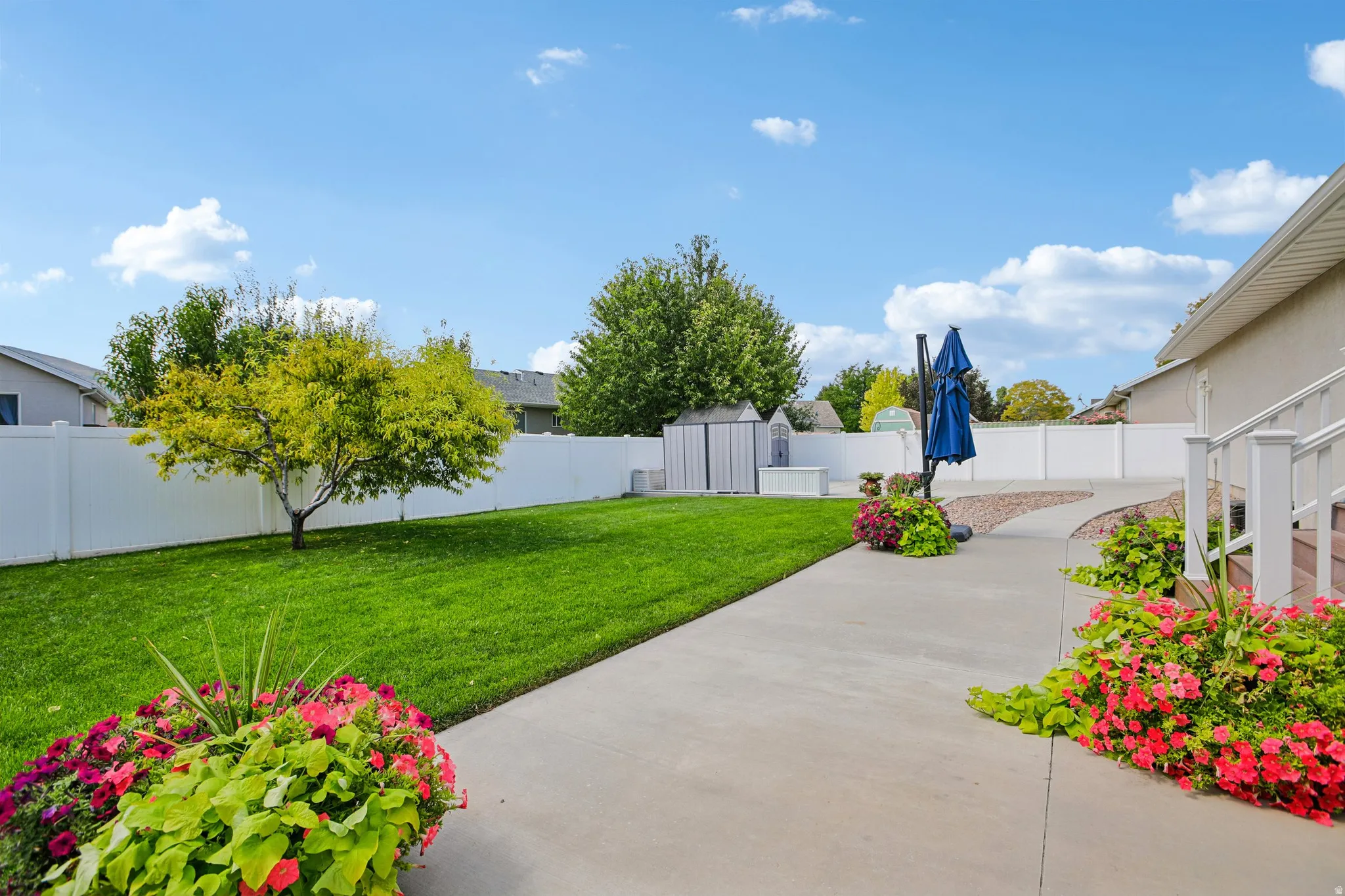 Expansive grassy yard with mature tree, white privacy fencing, concrete pathway, and vibrant landscaping