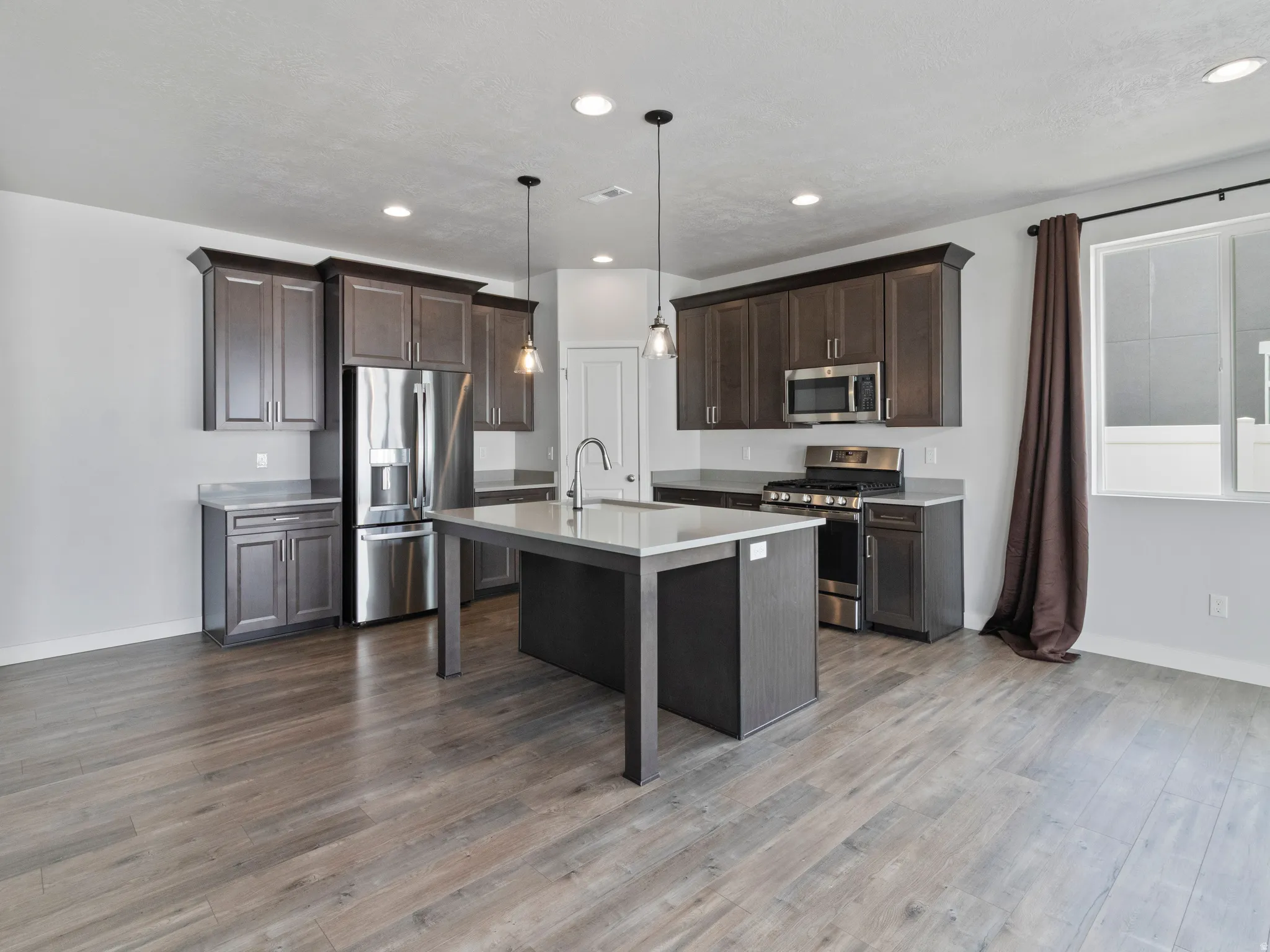 Chef's kitchen featuring dark cabinetry, stainless steel appliances including a gas range and refrigerator, quartz countertops, a large center island with seating, pendant lighting, and wide-plank wood-finish flooring throughout the open-concept space